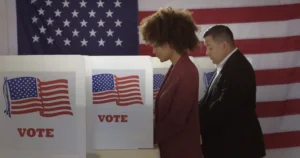 People in polling station, voting in a booth with US flag in background to illustrate Americans voting in the 2024 election. By vesperstock/stock.adobe.com