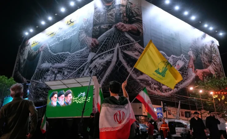 An Iranian demonstrator waves a flag of Lebanon's militant Hezbollah group under an anti-U.S. billboard depicting the American aircrafts into the Iranian armed forces fishing net with signs that read in Farsi: "The Strait of Hormuz will remain closed, The entire Persian Gulf is our hunting ground," during a pro-government gathering at Enqelab-e-Eslami, or Islamic Revolution, square in Tehran, Iran, Monday, May 4, 2026. (AP Photo/Vahid Salemi)