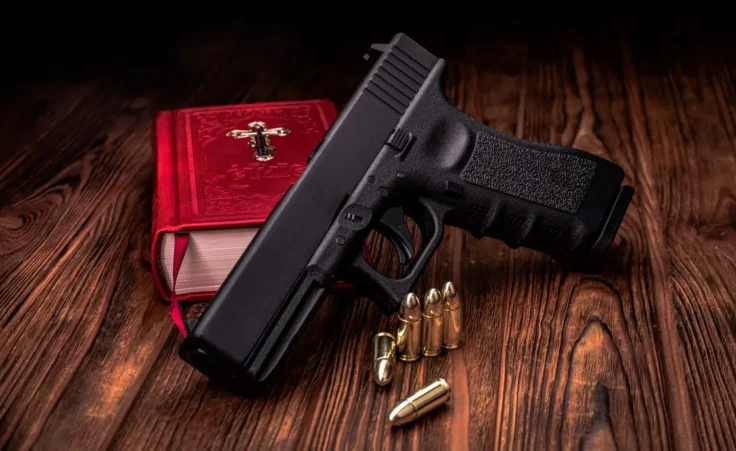 On a wooden table lies a pistol and cartridges, behind the pistol lies a bible in a red cover, soft focus By Евгений Мандажи/stock.adobe.com