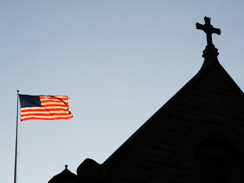 A cross atop a church with American flag below in the foreground (Credit: surpasspro via Fotolia.com)