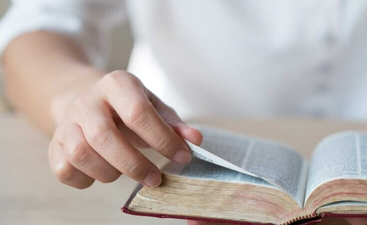 A person turns the page of an open Bible in their hand. © By manusapon/stock.adobe.com