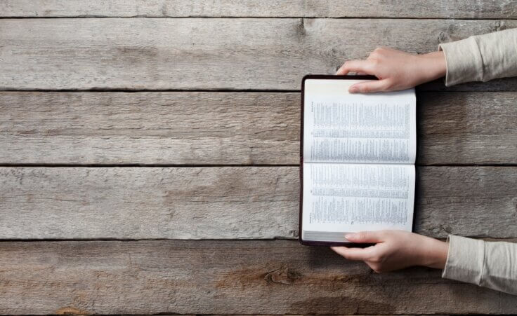 A woman holds a Bible open on a wooden table. © By 4Max/stock.adobe.com