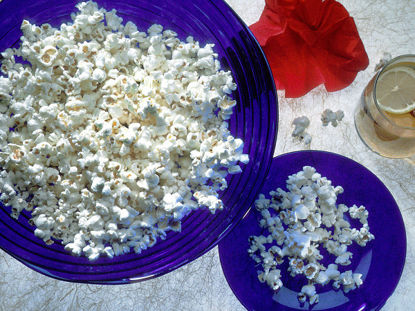There are royal blue bowls (1 large, 1 small) full of white popcorn, on a white tablecloth. Behind the bowls are a glass mug of tea with a lemon slice and a cinnamon stick and a red poppy (Credit: National Institutes of Health/National Cancer Institute)