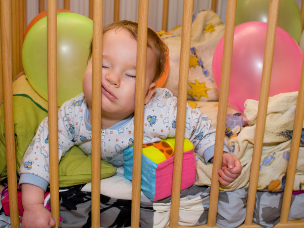 A cute little baby in pajamas asleep in crib with face pressed against the crib rails (Credit: st-fotograf via Fotolia.com)
