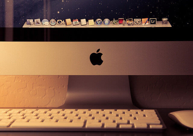 The iMac 2012 sitting on a desk in a softly lit office, with a closeup of the Apple logo (Credit: Manny R via Flickr)