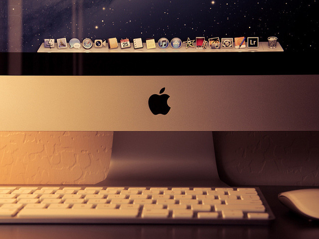 The iMac 2012 sitting on a desk in a softly lit office, with a closeup of the Apple logo (Credit: Manny R via Flickr)