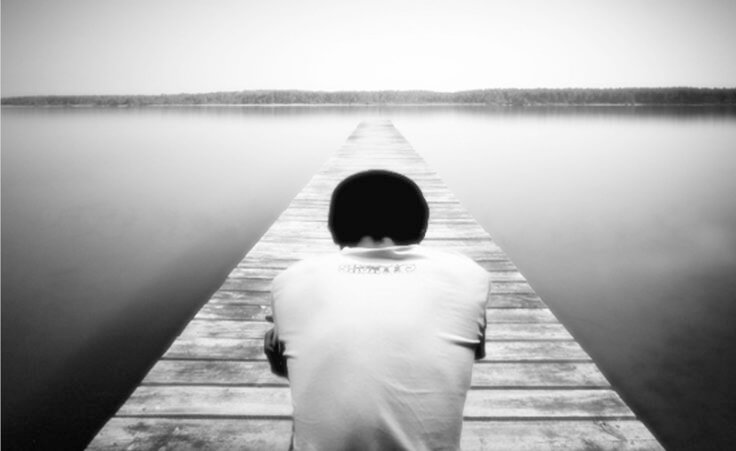 A boy sits alone on a dock that stretches into a fog shrouded lake (Credit: buaiansayapanomali via DeviantArt)