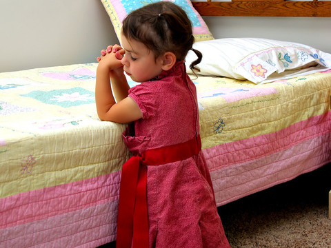 A young child, five years old maybe, kneeling beside her bed in her room, praying (Credit: Derek Miller via Fotolia)