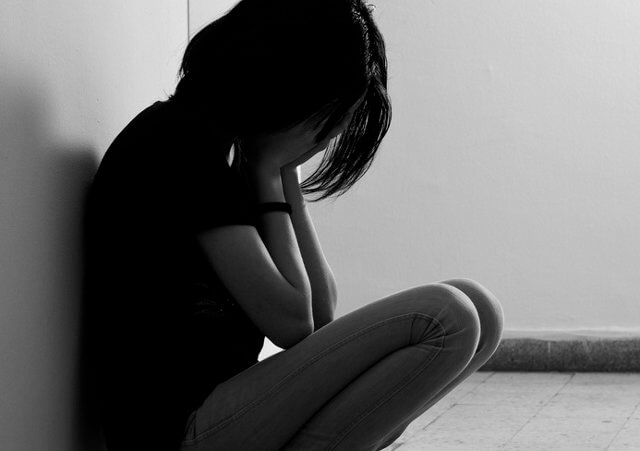 A depressed teenage girl with her face in her hands squatting on the floor with her back against a wall (Credit: Mitarart via Fotolia)