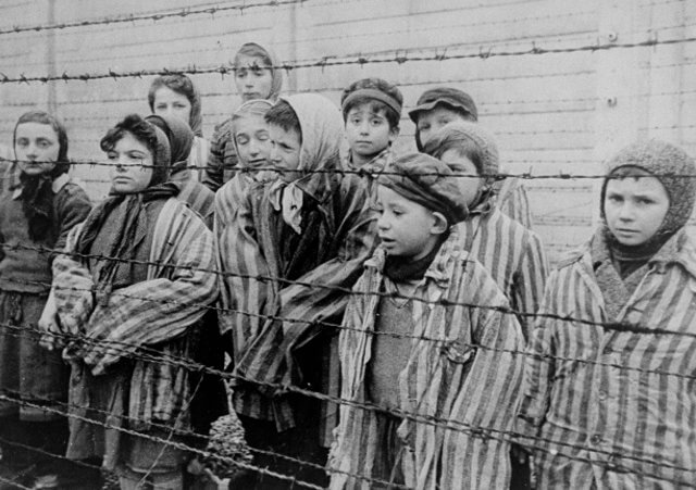 Child survivors of Auschwitz, wearing adult-size prisoner jackets, stand behind a barbed wire fence. Among those pictured are Tomasz Szwarz; Alicja Gruenbaum; Solomon Rozalin; Gita Sztrauss; Wiera Sadler; Marta Wiess; Boro Eksztein; Josef Rozenwaser; Rafael Szlezinger; Gabriel Nejman; Gugiel Appelbaum; Mark Berkowitz (a twin); Pesa Balter; Rut Muszkies (later Webber); Miriam Friedman; and twins Miriam Mozes and Eva Mozes wearing knitted hats (Credit: USHMM/Belarusian State Archive of Documentary Film and Photography/Alexander Voronzow and others in his group, ordered by Mikhael Oschurkow, head of the photography unit)