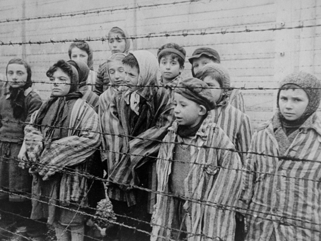 Child survivors of Auschwitz, wearing adult-size prisoner jackets, stand behind a barbed wire fence. Among those pictured are Tomasz Szwarz; Alicja Gruenbaum; Solomon Rozalin; Gita Sztrauss; Wiera Sadler; Marta Wiess; Boro Eksztein; Josef Rozenwaser; Rafael Szlezinger; Gabriel Nejman; Gugiel Appelbaum; Mark Berkowitz (a twin); Pesa Balter; Rut Muszkies (later Webber); Miriam Friedman; and twins Miriam Mozes and Eva Mozes wearing knitted hats (Credit: USHMM/Belarusian State Archive of Documentary Film and Photography/Alexander Voronzow and others in his group, ordered by Mikhael Oschurkow, head of the photography unit)