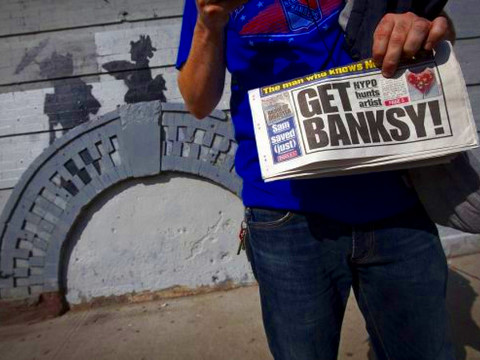 A man holds up a local newspaper as he poses for a photo in front of a new art piece by British graffiti artist Banksy in the Brooklyn borough of New York, October 17, 2013. (Credit: Reuters/Carlo Allegri)