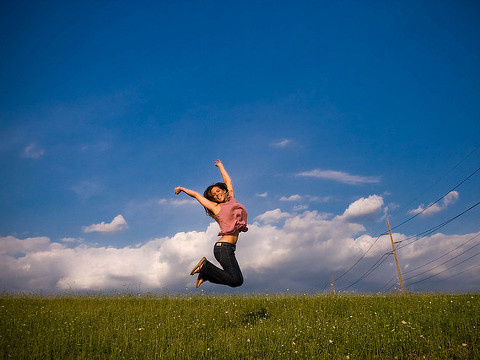 A young woman jumping in the air with joy in a field of green against a bright blue sky (Credit: Marcy Kellar via Flickr)