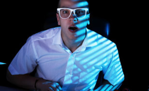 A young man in a white shirt and glasses stares in shock at his computer screen while browsing the internet (Credit: Yeko Photo Studio via Fotolia)