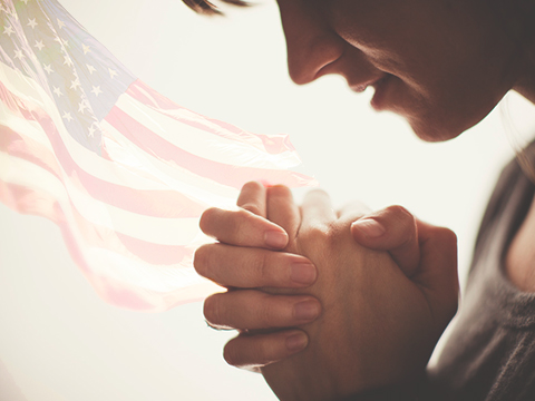 A young woman with her hands folded in prayer against a muted American flag background (Credit: Evan Burton using works from Chad Madden and Shaun Menary via Lightsock)