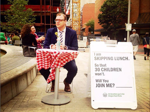 Portland native, Anton Cobb, on the first day of his campaign to raise money for the local food bank, sits at small table with a red-checkered tablecloth in Director's Park, with a sign that reads 'I am skipping lunch. So that 30 children won't. Will you join me? Donate your lunch money to the Oregon Food Bank', July 24, 2014 (Credit: Heather Boyd via hOURLUNCH on Facebook)