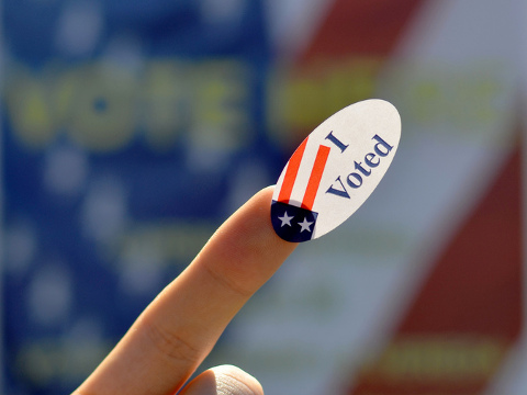 A voter holds an 'I Voted' sticker on index finger in front of 'Vote Here' sign outside a polling location in Berkeley, California, November 6, 2012 (Credit: Daniel Parks via Flickr)