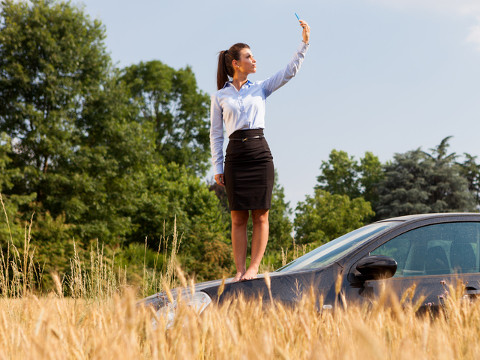 A young Italian businesswoman out in the country searches for a cell phone signal while standing on the hood of her car (Credit: Francesco83 via Fotolia)