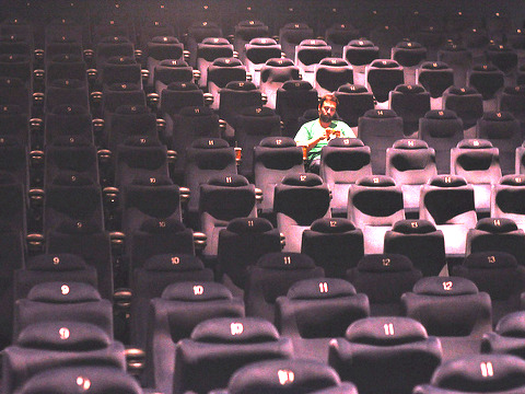 Maegan Tintari's husband sits in theater along looking at a phone or maybe a box of candy before the start of the movie Take Me Home Tonight (Credit: Maegan Tintari via Flickr)
