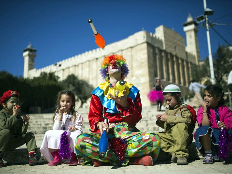 A Jewish settler dressed as a clown juggles next to children as the Tomb of the Patriarchs is seen in the background, during a parade marking the Jewish holiday of Purim in the West Bank city of Hebron, March 5, 2015 (Credit: Reuters/Amir Cohen)