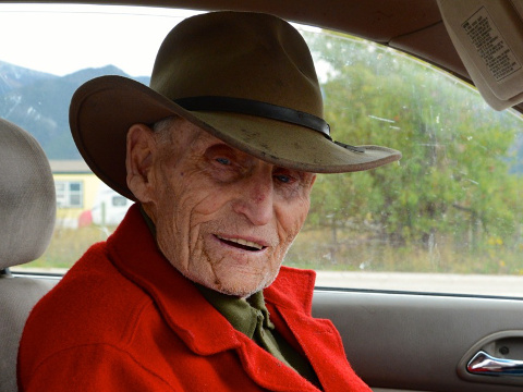 Lloyd Baker, wearing a red flannel coat, sits in his car, outside the cabin where he was born 104 year ago in Etna, Wyoming (Credit: National Geographic Traveler/Andrew Evans)