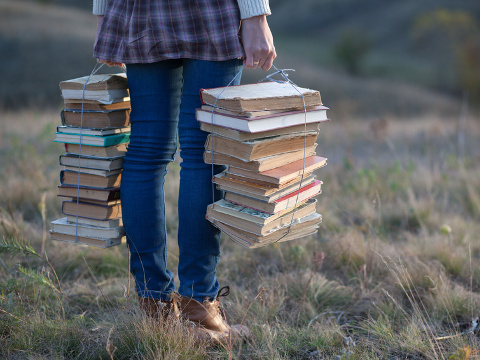 Hipster girl holding two stacks of books, tied with cord, in an open field in autumn (Credit: rasstock via Fotolia)