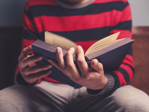 Man in gray sweats and a red-and-blue striped sweater sitting on a bench is reading a big book (Credit: LoloStock via Fotolia)
