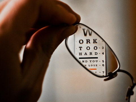 Work Too Hard printed on an eye exam chart viewed through a single eyeglass lens being held by a patient (Credit: Chris Martino via Flickr)