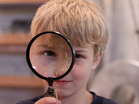 A young, preteen, blond-haired boy looking through a magnifying glass playing junior detective (Credit: Jessica Lucia via Flickr)