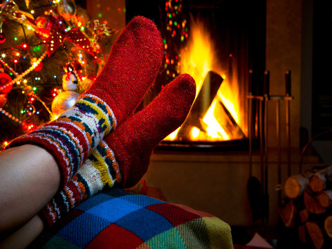 A woman with stripped socks sitting in a chair by the fireplace and her lit and decorated Christmas tree (Credit: Konstiantyn via Fotolia)