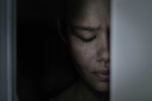 Half of a sad woman's face, her eyes closed in distress, appears behind a doorframe. By globalmoments/stock.adobe.com
