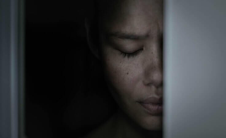 Half of a sad woman's face, her eyes closed in distress, appears behind a doorframe. By globalmoments/stock.adobe.com