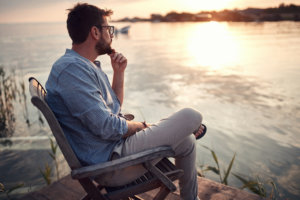 A man sits deep in thought next to a lake with his left hand resting on his chin, possibly asking himself, "Why should I believe in God?" © luckybusiness /stock.adobe.com