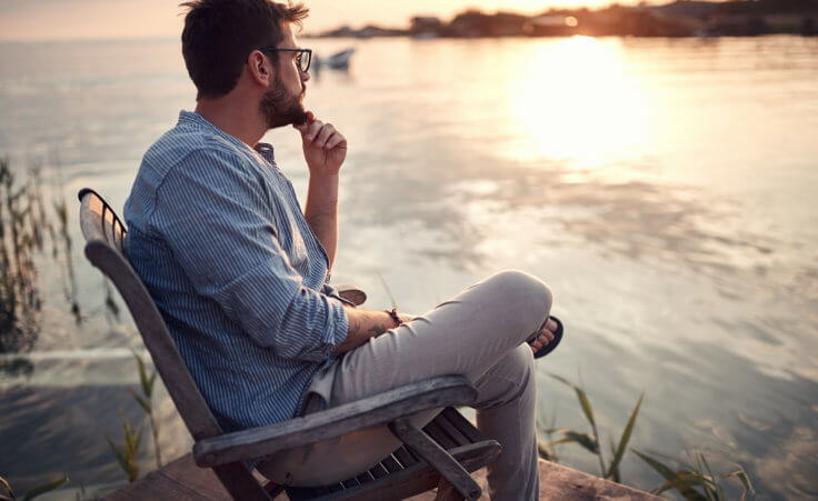 A man sits deep in thought next to a lake with his left hand resting on his chin, possibly asking himself, "Why should I believe in God?" © luckybusiness /stock.adobe.com