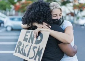 Two women hug, one has an "End Racism" banner in her hands. By Sabrina/stock.adobe.com
