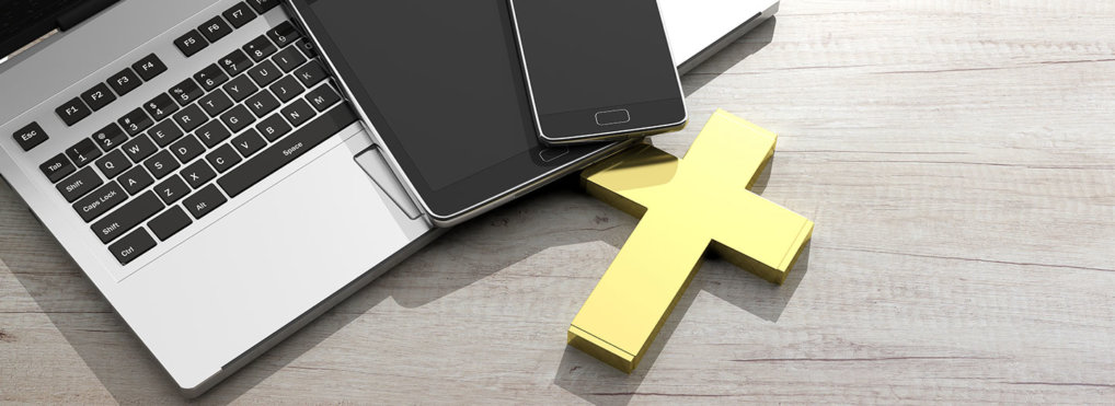 A yellow cross, MacBook, iPad, and iPhone sit on a wooden desk.