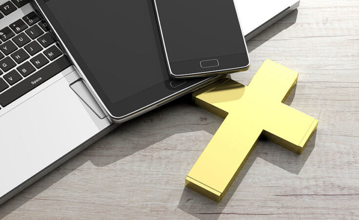 A yellow cross, MacBook, iPad, and iPhone sit on a wooden desk.