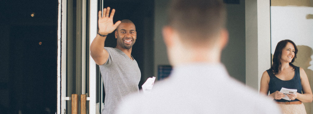 A man standing in front of open church doors waves to an oncoming visitor.