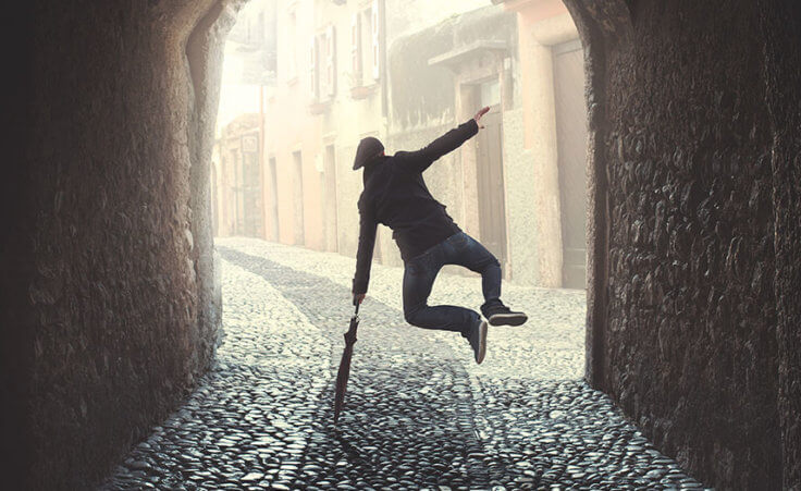 Inside a dark tunnel, a man leaps into the air and clicks his heels while balancing on a closed umbrella