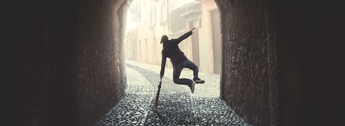 Inside a dark tunnel, a man leaps into the air and clicks his heels while balancing on a closed umbrella