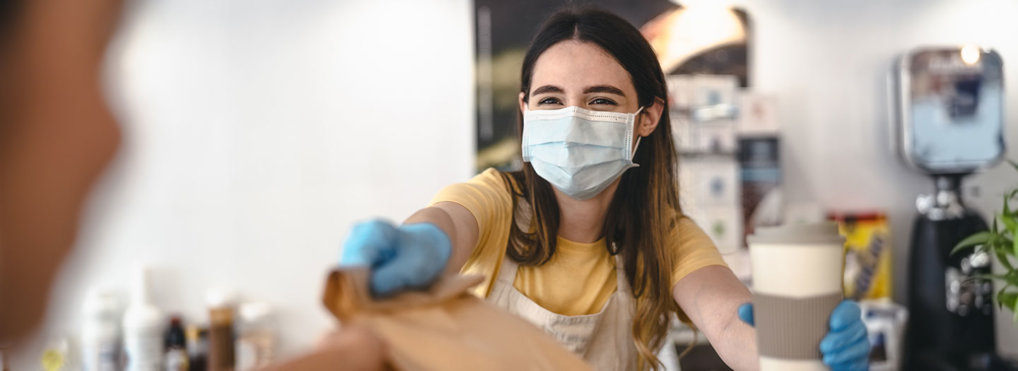 A female barista in a face mask hands a paper sack and a cup of coffee to a customer