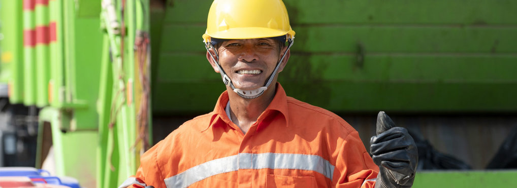 A garbage man dressed in an orange jumpsuit smiles while giving a thumbs up in front of a green garbage truck