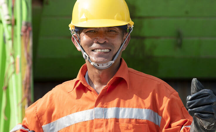 A garbage man dressed in an orange jumpsuit smiles while giving a thumbs up in front of a green garbage truck