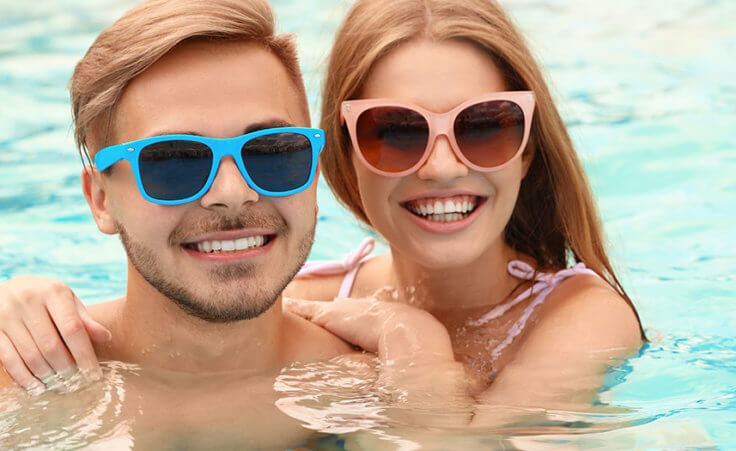 A man and woman, both wearing sunglasses, float in a pool while smiling for the camera