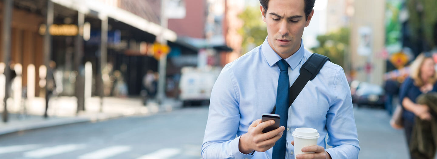 A young professional stares at his phone while holding his coffee and walking down the street