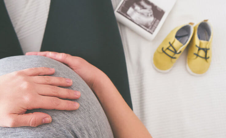Seen from above, a pregnant woman covers her stomach with her hand. Baby shoes and ultrasound photos lay next to her.