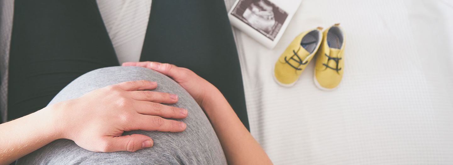 Seen from above, a pregnant woman covers her stomach with her hand. Baby shoes and ultrasound photos lay next to her.