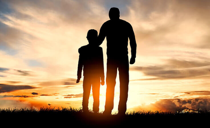 A father and child stand in silhouette, the sun in the distance, the father's hand over the child's shoulders