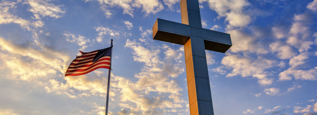The American flag waves next to a cross against a cloudy blue sky.