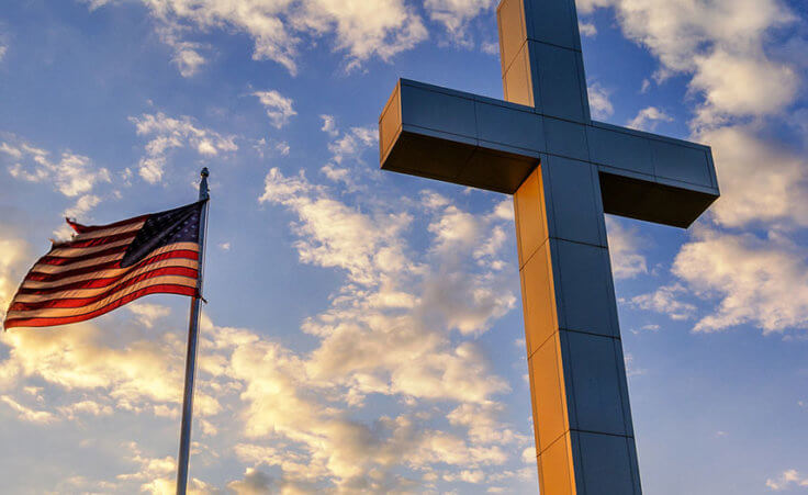 The American flag waves next to a cross against a cloudy blue sky.